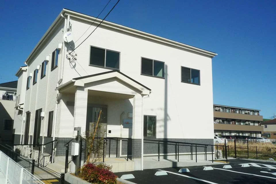 ブルーミングケアの白い2階建て建物の外観で、車椅子用スロープと駐車場が整備されています。